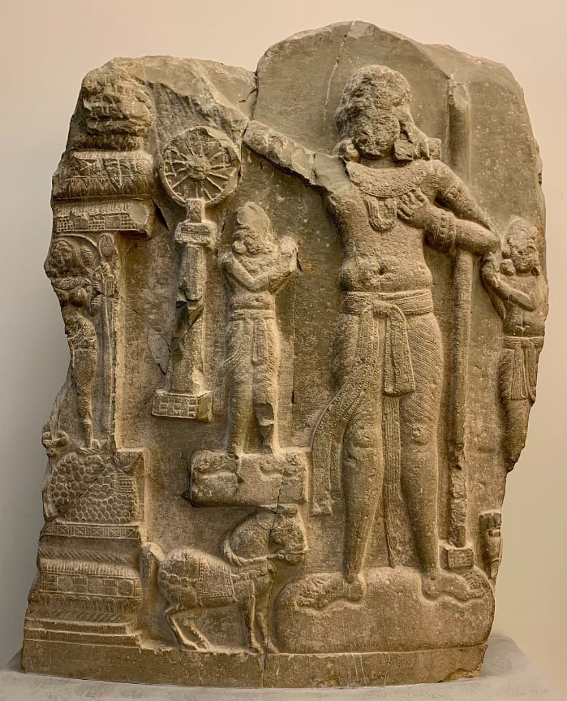 Ancient stone relief carving depicting Emperor Ashoka gesturing toward the Dharmachakra on a pillar
